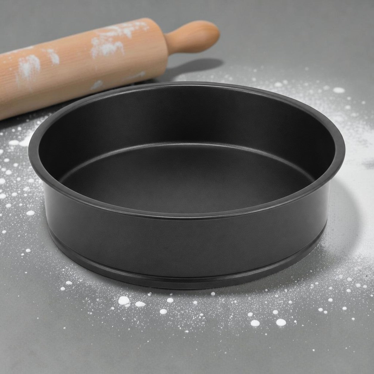 Tap2Own 9-inch round black nonstick cake pan made of heavy-duty steel with a rolling pin in the background.
