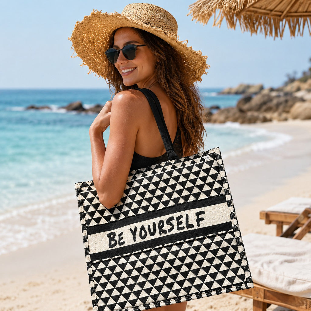 Tap2Own Be Yourself graphic tote bag, black and white geometric pattern, slogan across center, black handles, beach setting