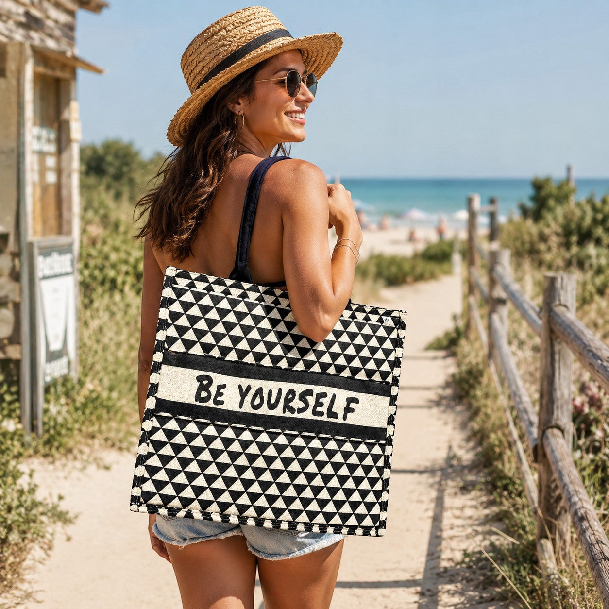 Tap2Own Be Yourself graphic tote bag with black and white geometric pattern and black handles, carried by a woman on a beach path.