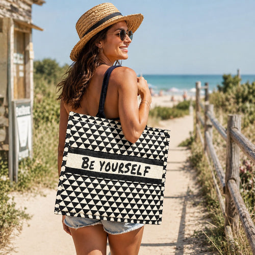 Tap2Own Be Yourself graphic tote bag with black and white geometric pattern and black handles, carried by a woman on a beach path.