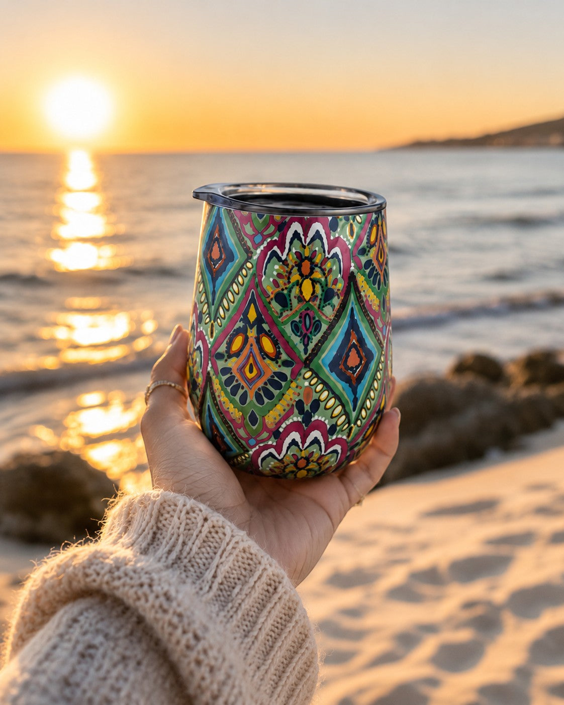 Tap2Own stainless steel insulated wine tumbler with a colorful bohemian pattern held on a beach at sunset.