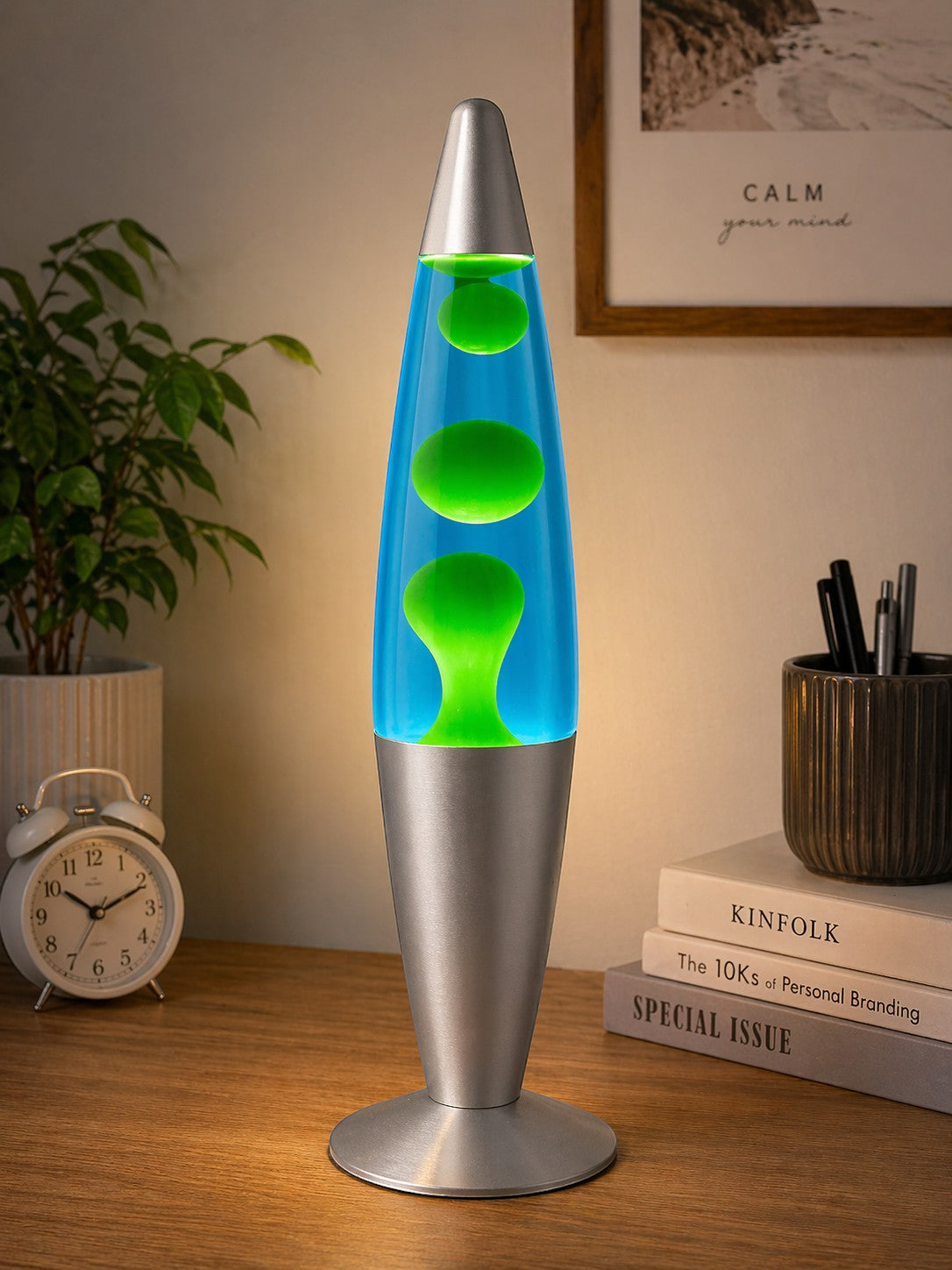 Tap2Own retro lava lamp with blue liquid and green blobs, silver base and cap, on a wooden desk.